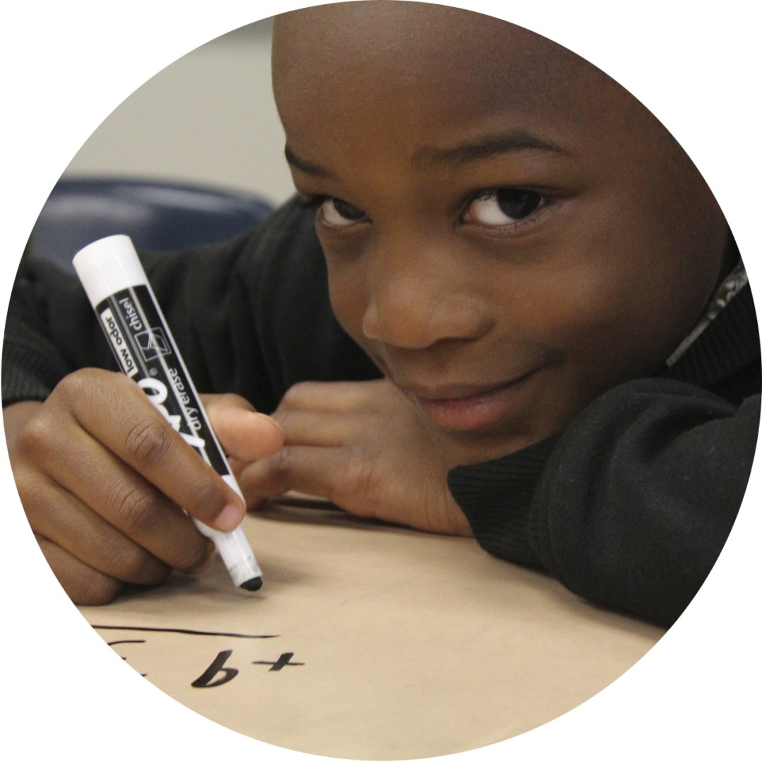A young child, smiling softly to the camera, holds a marker to paper, writing a math equation.